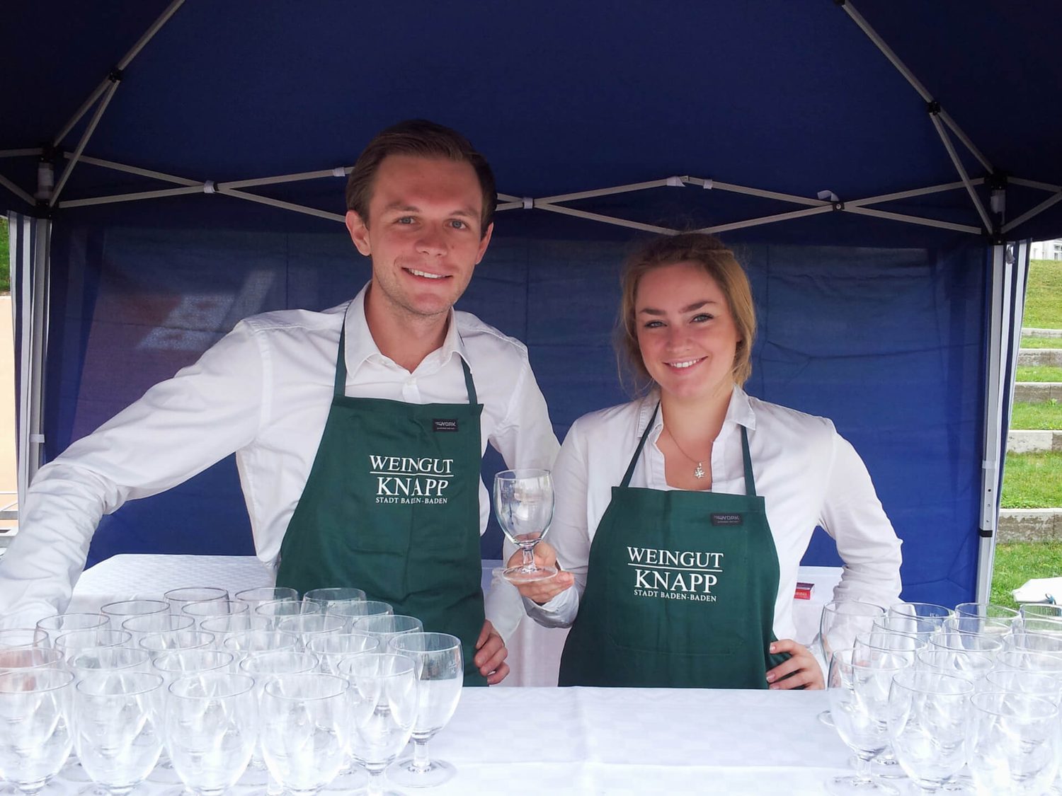 Zwei Mitarbeiter des Weinguts Knapp in gebrandeten grünen Schürzen am Weinprobe-Stand